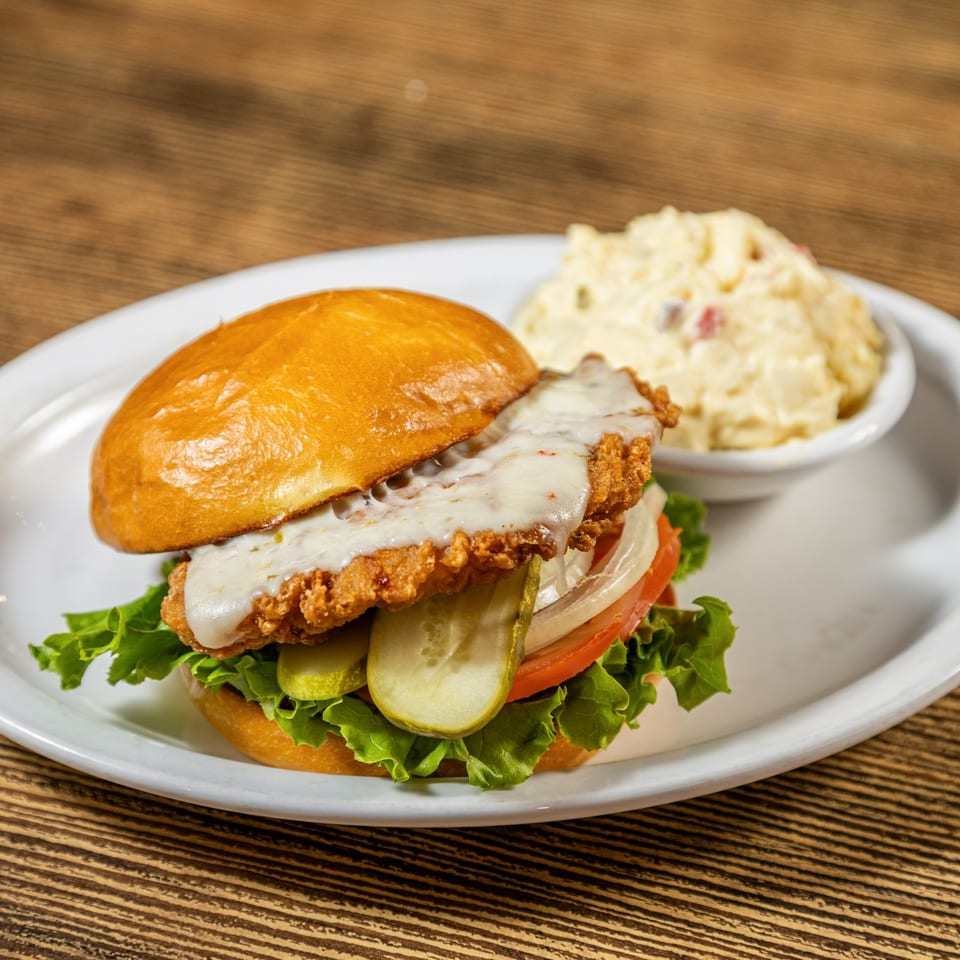 Crispy chicken sandwich with melted cheese, lettuce, tomato, onion, pickles, and a side of potato salad.
