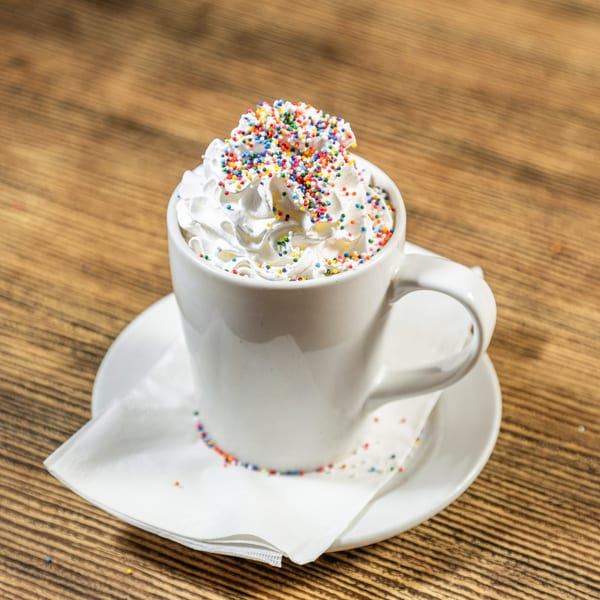 White mug of hot chocolate topped with whipped cream and colorful sprinkles on a wooden table.