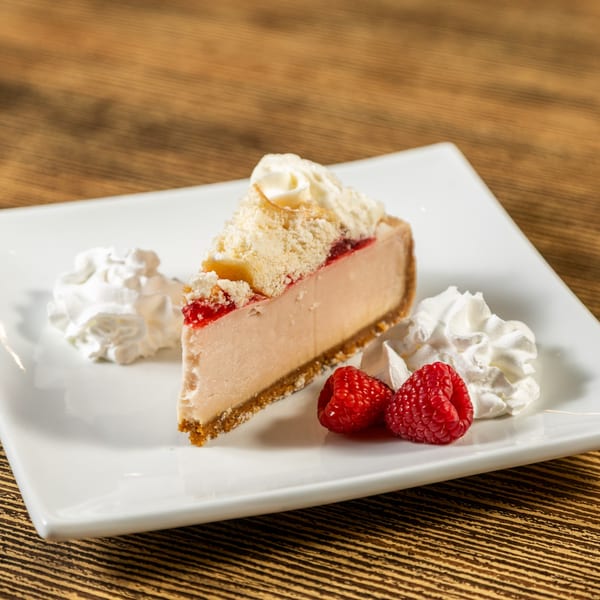 Slice of cheesecake with raspberry topping, whipped cream, and fresh raspberries on a white plate.