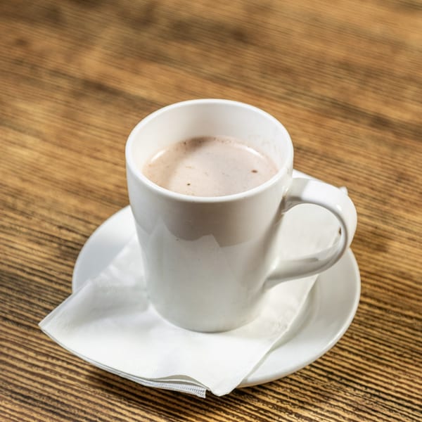 A white mug of hot chocolate with foam sits on a saucer with a napkin on a wooden table.