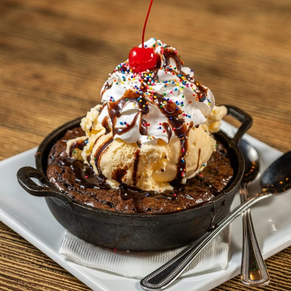 Warm brownie skillet topped with vanilla ice cream, whipped cream, sprinkles, chocolate syrup, and a cherry.