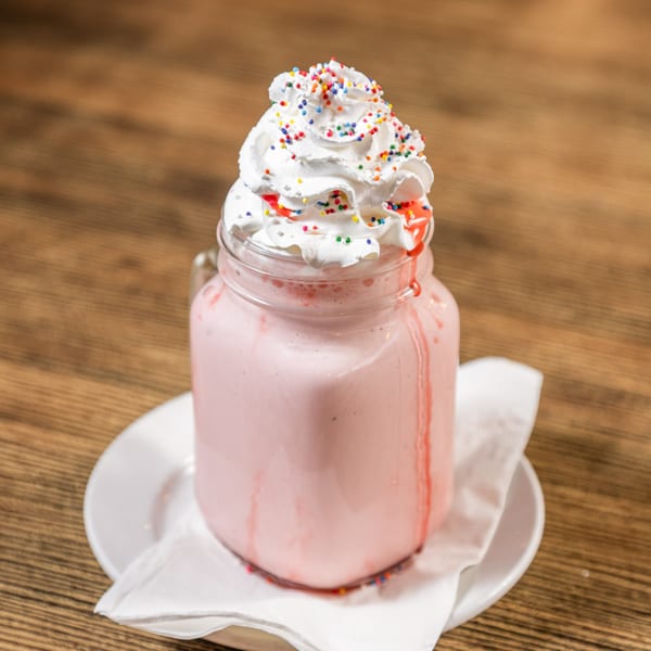 Pink milkshake in a mason jar, topped with whipped cream and colorful sprinkles, on a wooden table.