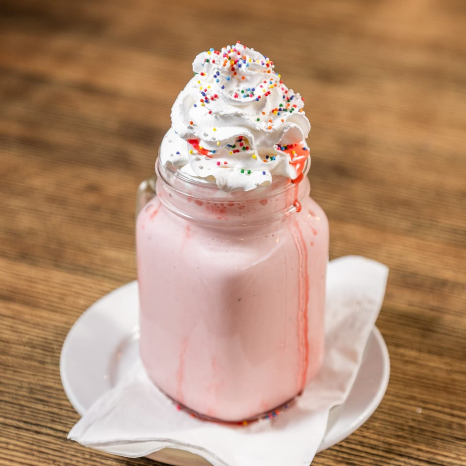 Pink milkshake in a mason jar, topped with whipped cream and colorful sprinkles, on a wooden table.